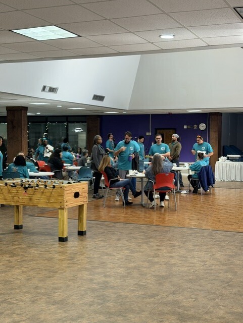 Attendees wearing matching teal "Students First" T-shirts sit in rows of chairs facing a stage during a back-to-school welcome event. A speaker presents at a podium while presentation screens display event content at the front of the room. Colorful balloon decorations line the stage area, and a large inflatable welcome display is visible to the left. The event takes place in a spacious meeting hall with high ceilings and bright overhead lighting.