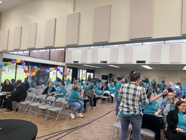 A large group of school employees wearing matching teal "Students First" T-shirts gathers in a meeting hall during a back-to-school welcome event. Attendees are seated in rows of folding chairs and conversing before the program begins. A colorful balloon arch decorates the entrance area, and large windows along one wall provide natural light to the room.