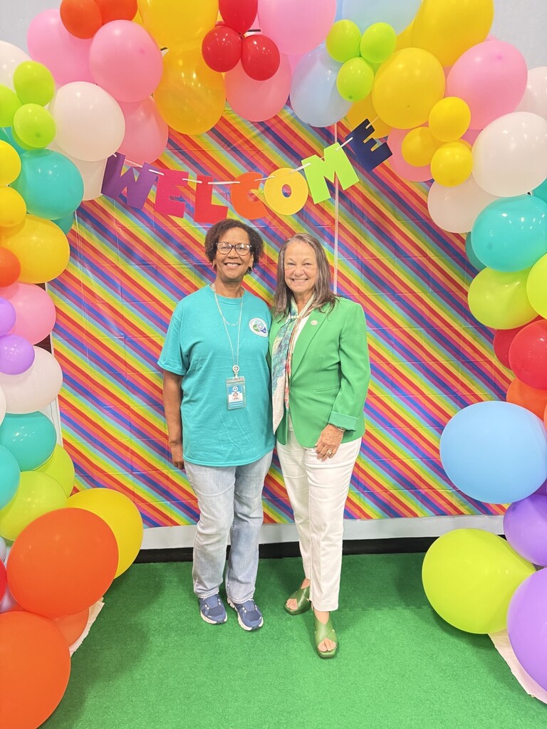 Dr. Shnirley Lewis and Sandy Hollaway pose for a photo in front of a colorful back-to-school display featuring a rainbow-striped backdrop, a "WELCOME" banner, and a large balloon arch made of pink, yellow, green, blue, orange, purple, and white balloons. One person wears a teal event T-shirt and identification badge, while the other wears a green blazer and light-colored pants. The pair stands on a green carpet beneath the festive decorations.