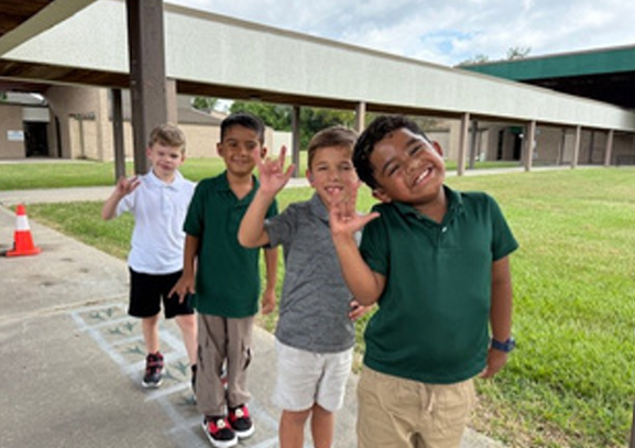 Four LSD students stand on a covered school walkway outside a campus building. The students are facing the camera with one hand raised, while grassy areas, concrete sidewalks, and school buildings are visible in the background.