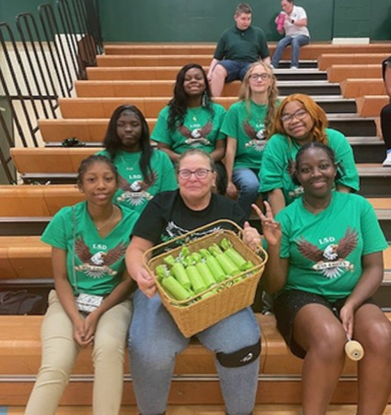 Amy Jarrett sits with a group of students on gymnasium bleachers wearing matching green school shirts featuring an eagle design. The adult in the front row holds a basket filled with wrapped green items, while one student nearby holds up a hand gesture. Additional individuals are seated on the bleachers in the background.