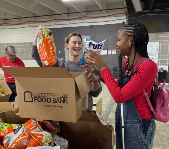 olunteers sort donated food items into boxes at the Greater Baton Rouge Food Bank warehouse. One volunteer holds a bag of vegetables while another holds a package of paper towels over an open donation box filled with food products.