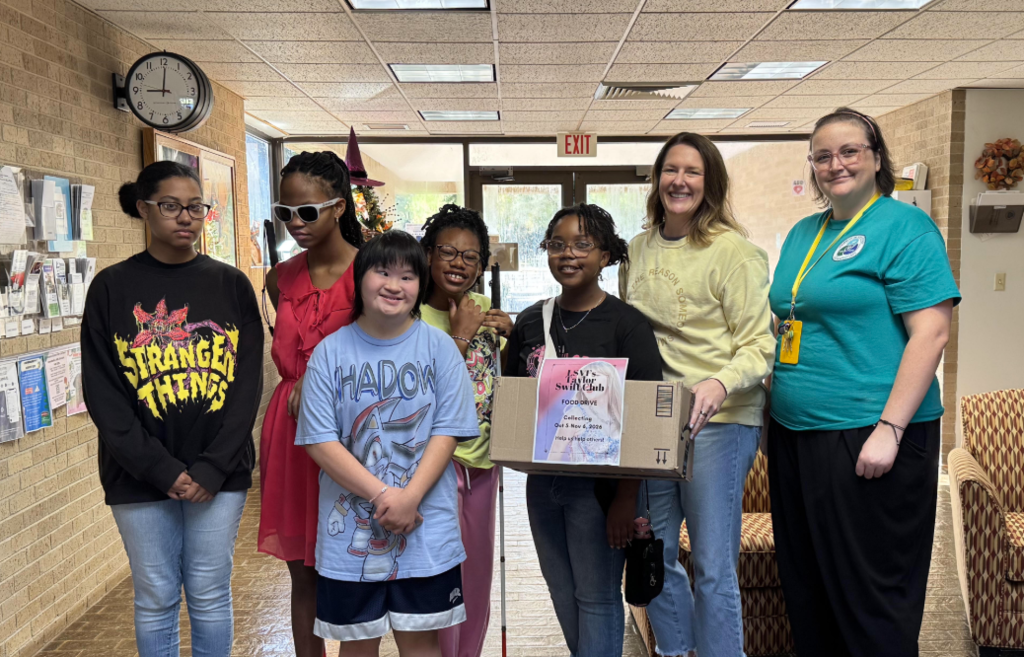 A group of students and staff members pose indoors with a donation box for a food drive. The box is labeled for a school club food drive, highlighting a community service project supporting local food assistance efforts.