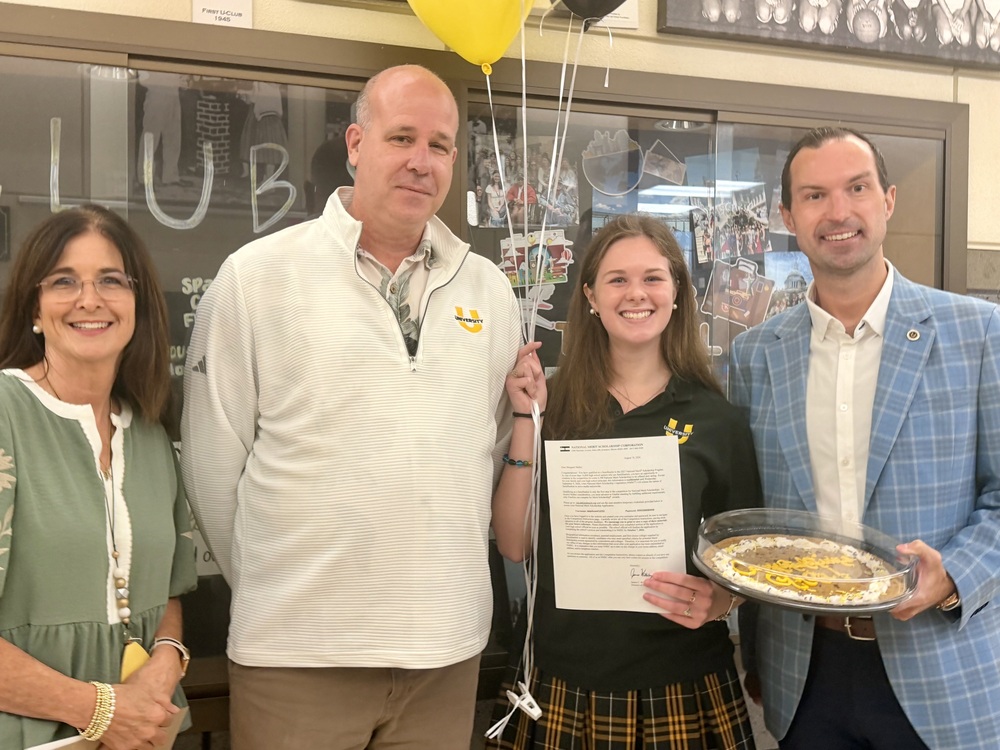 A student proudly holding a National Merit Scholarship letter poses with three adults holding a decorated pie and balloons.