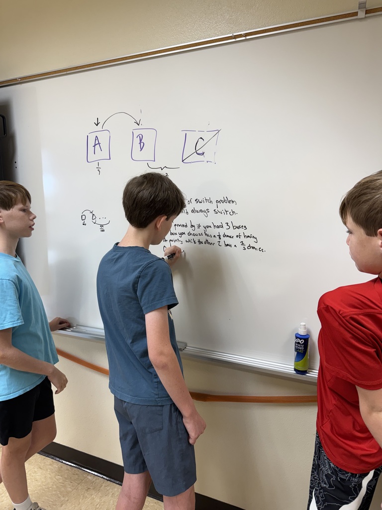 three boys in light colored shirts facing dry erase board working on math problems