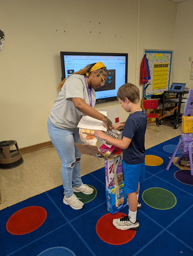 Female college student assisting a young boy with a cardboard project standing on a blue carpet in the classroom with a tv monitor on