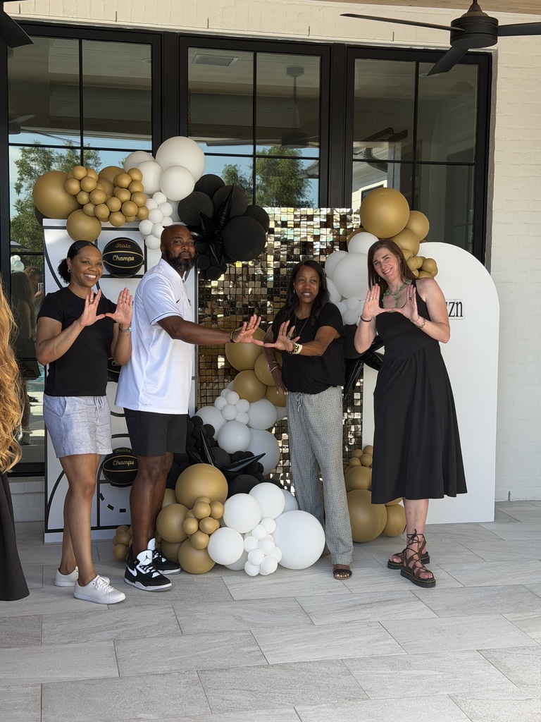 A small group of school administrators, gathers outside for a group photo in front of black, white, and gold balloon decorations. The coaches smile and proudly hold up their hands to display "U", celebrating this milestone together.