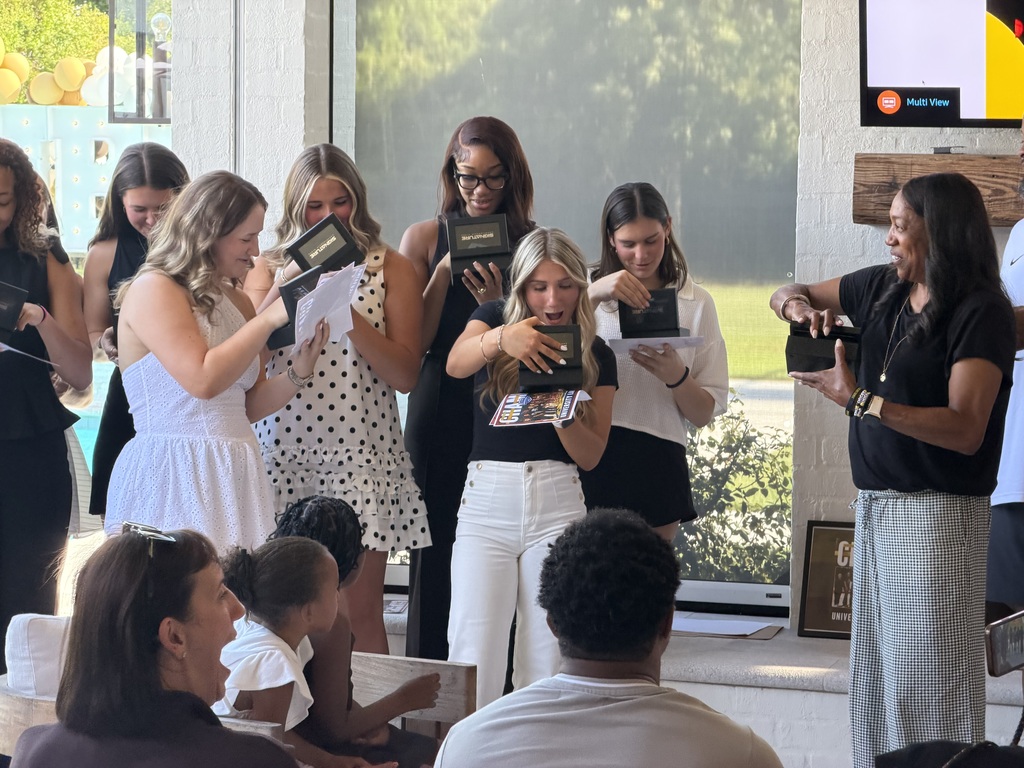 Students react with excitement and surprise as they open black ring boxes during a senior ring ceremony. Surrounded by classmates, family members, and school staff, they smile and admire their new senior rings while sharing the special moment together.