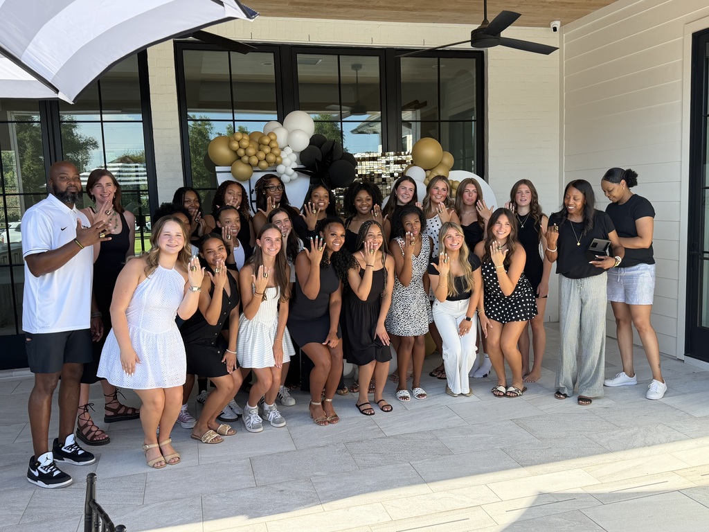 A large group of high school seniors, joined by school administrators, gathers outside for a group photo in front of black, white, and gold balloon decorations. The students smile and proudly hold up their hands to display their senior rings, celebrating this milestone together.