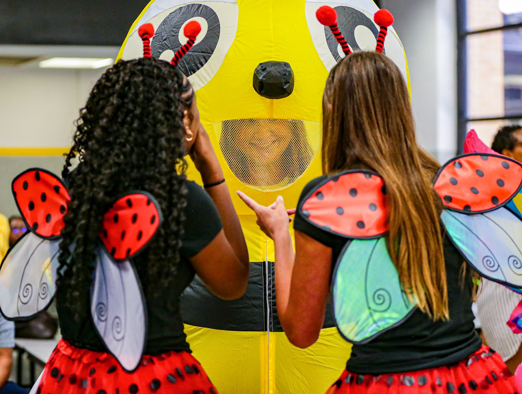 Two people in colorful insect costumes face a large yellow bee costume indoors.