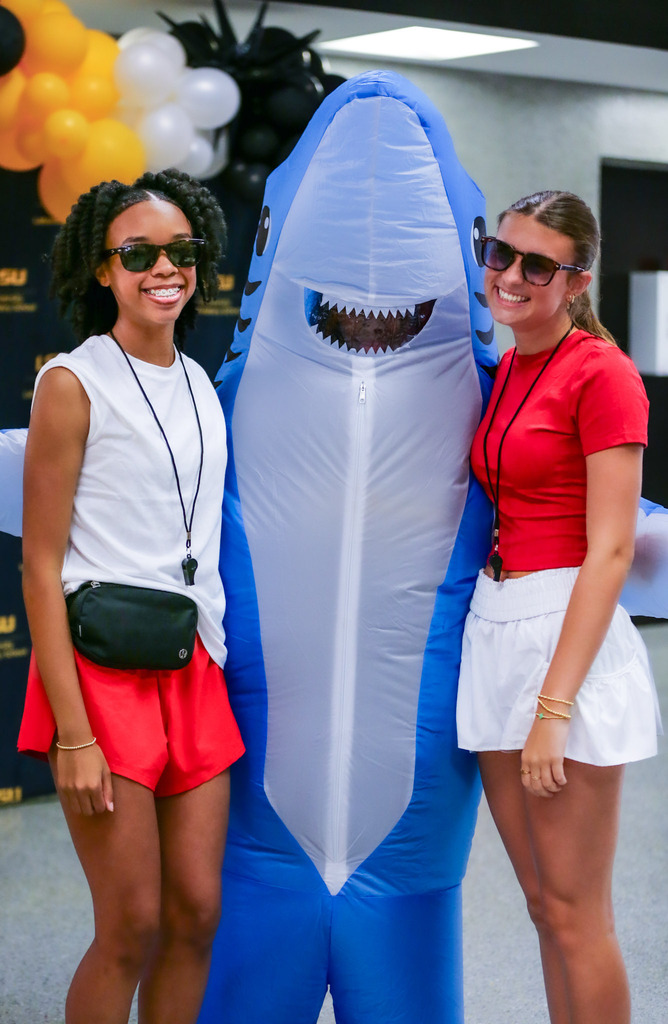 Two smiling women pose with a blue shark mascot at a team event.
