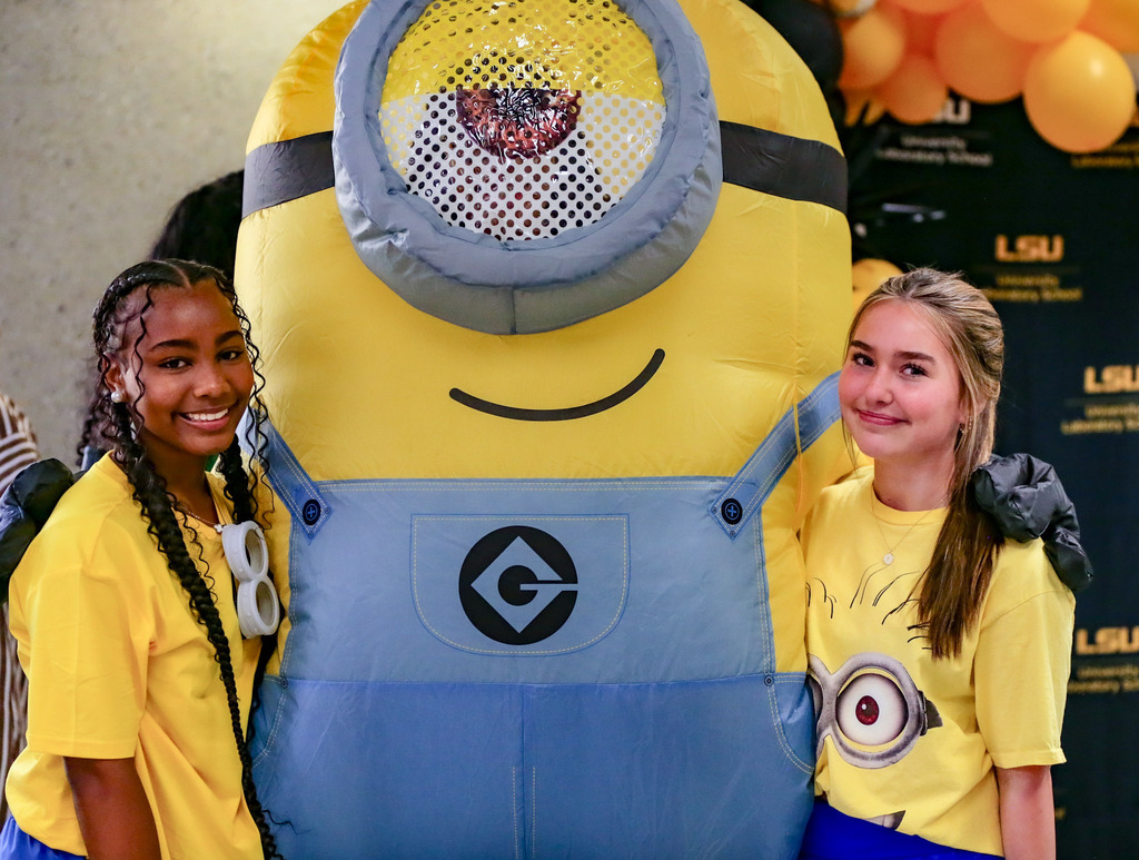 Two students in Minion-themed shirts pose with a large Minion mascot at a school event.