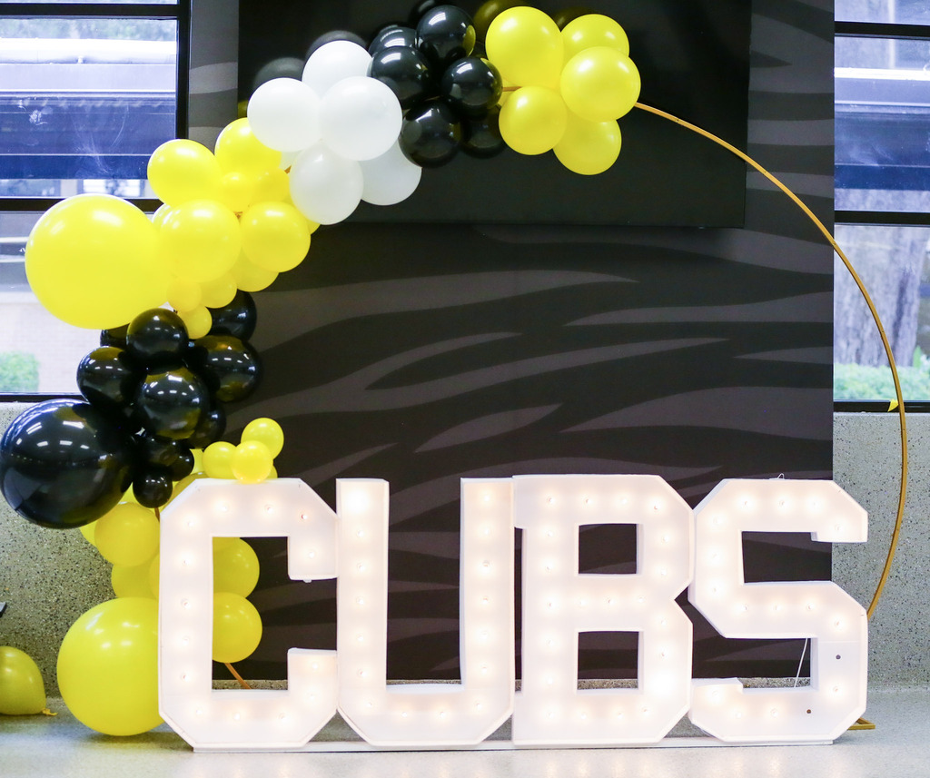 Large marquee letters spelling "CUBS" illuminated with fair lights sit in front of a dark backdrop with a subtle pattern, decorated with a balloon arch of yellow, black, and white balloons on a gold circular frame.