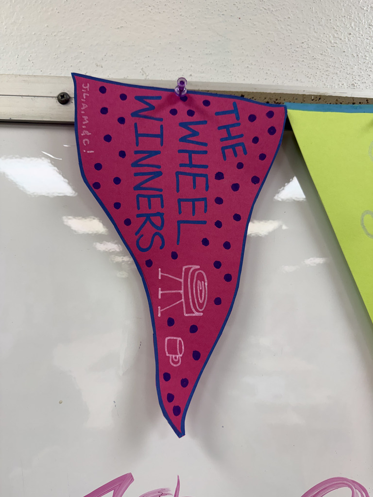 A pink paper pennant decorated with purple dots and blue text reading "THE WHEEL WINNERS," featuring a white illustration of a pottery wheel.