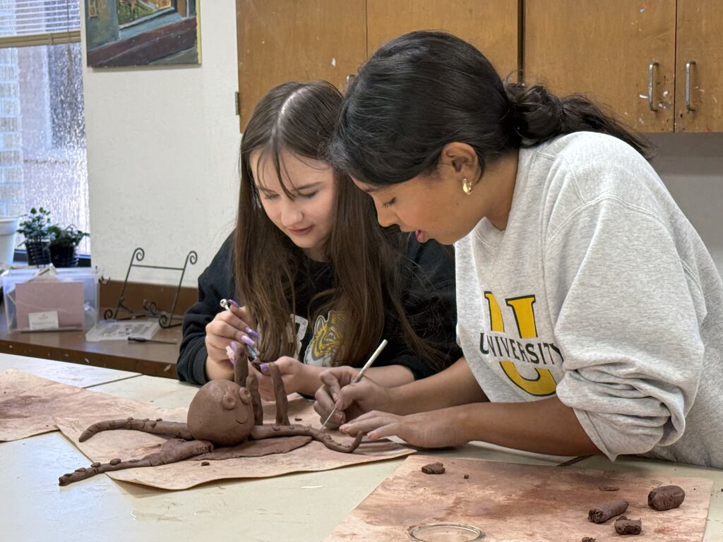 Two young women working together at a table to sculpt a detailed brown clay octopus using styling tools.