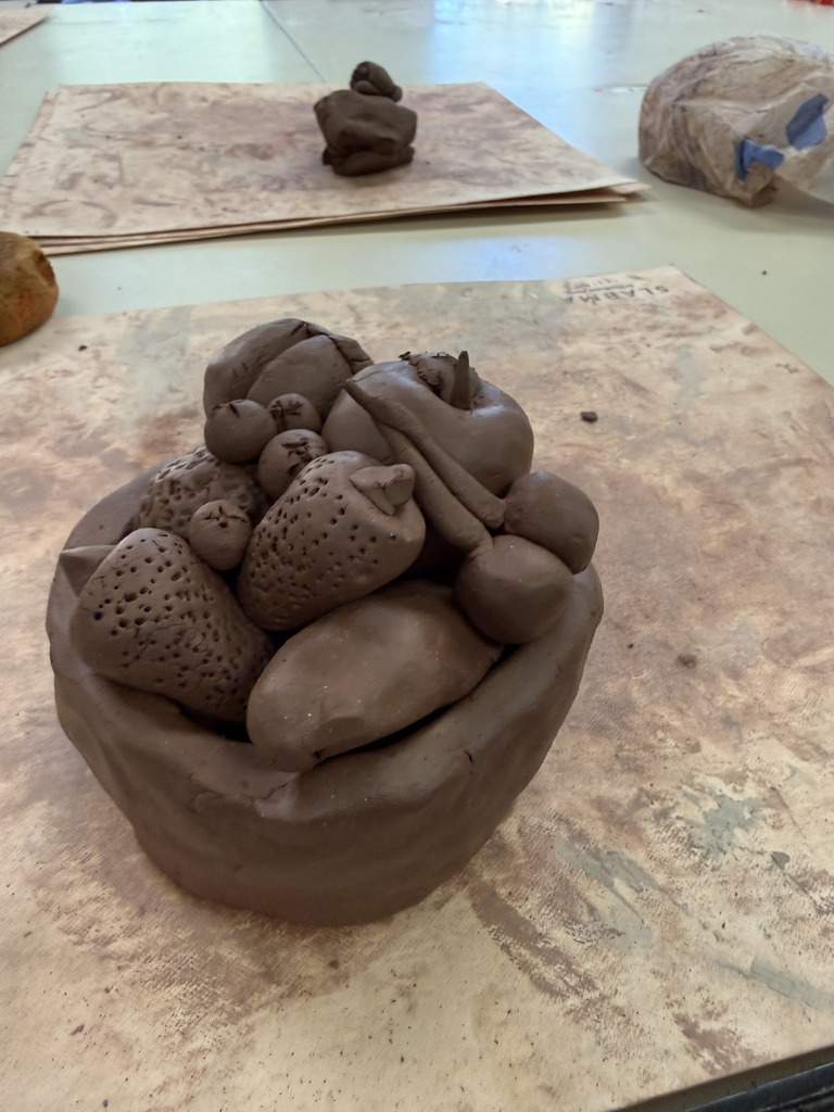 A dark brown clay sculpture of a bowl filled with various fruits, including strawberries, cherries, and a banana.