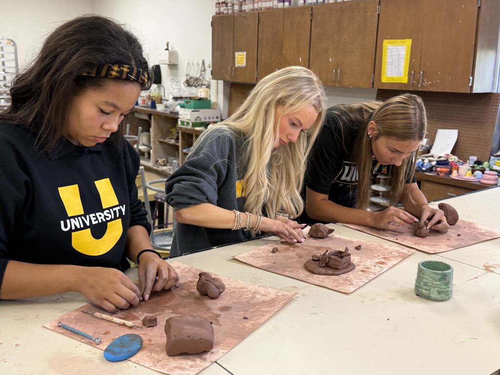 Three young women sitting at a table in an art classroom, focused on shaping brown clay on individual work mats.