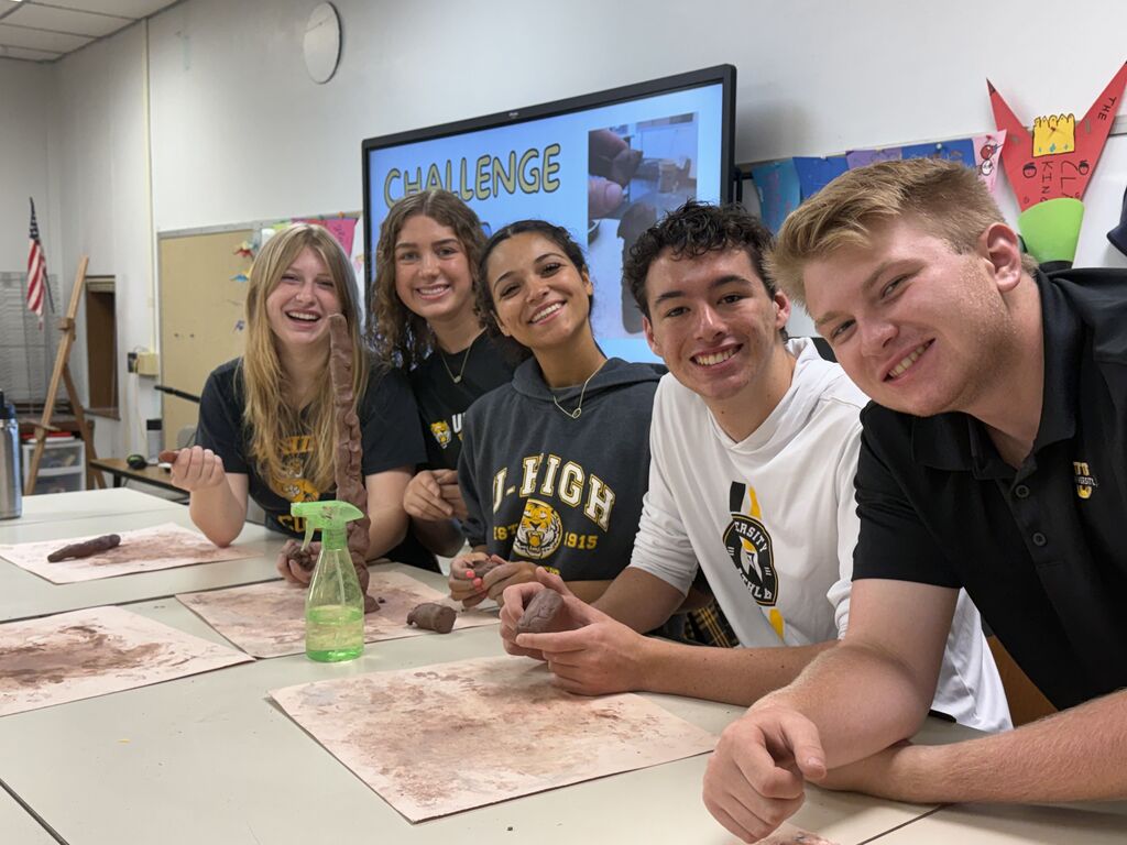 Five smiling teenagers posing together at a classroom table with their clay projects during a sculpting challenge.