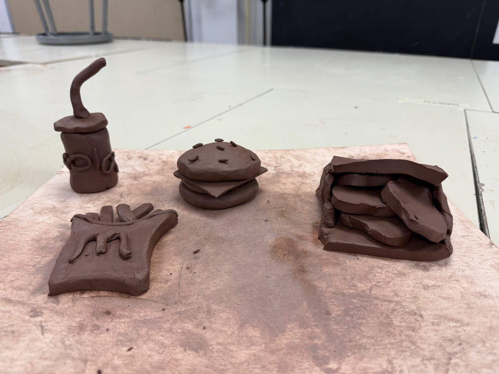 Four small brown clay sculptures representing a fast-food meal, including a burger, french fries, a drink with a straw, and a small box.
