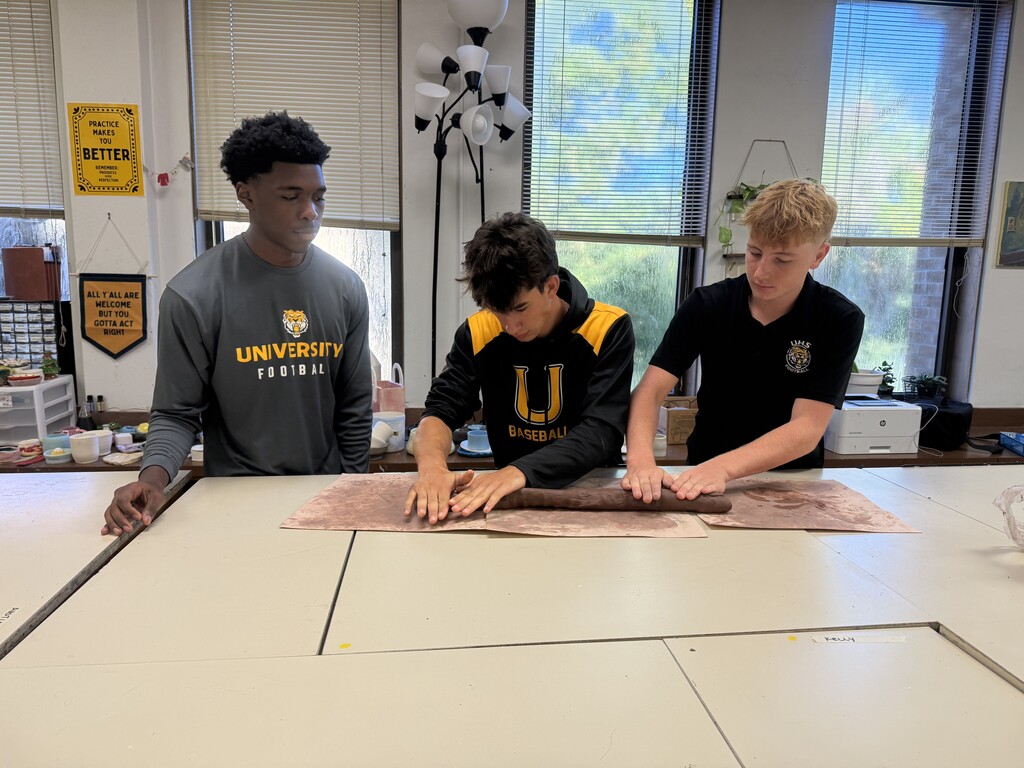 Three young men standing at a classroom table, working together to roll out a long log of brown clay.