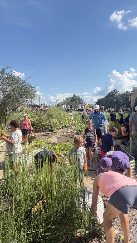 Children and adults tend to raised garden beds next to a football field where a team practices under a bright, cloudy sky.