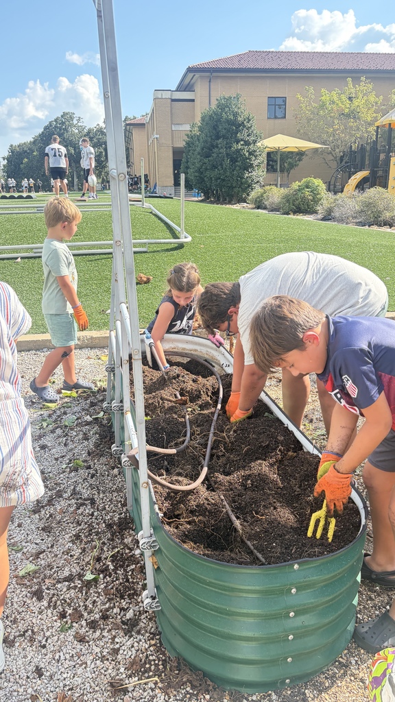 Children dig and work the soil inside a green, corrugated raised garden bed during an outdoor community activity near a school building.