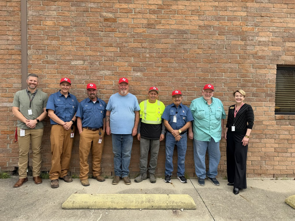 Eight people in red caps and casual work attire pose in front of a brick building.