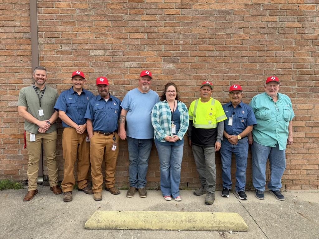Nine people stand in a row outside a brick building, wearing various colors of shirts and pants.