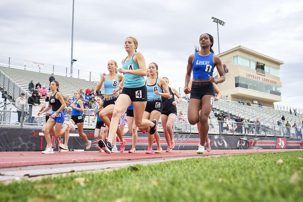 A group of female athletes compete in a track race. They wear matching uniforms. Spectators watch from the stands.