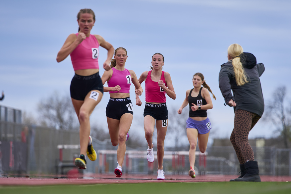 Five women sprint on a track, numbered from 2 to 8. They wear matching tops and shorts in various colors. Behind them, a woman watches.