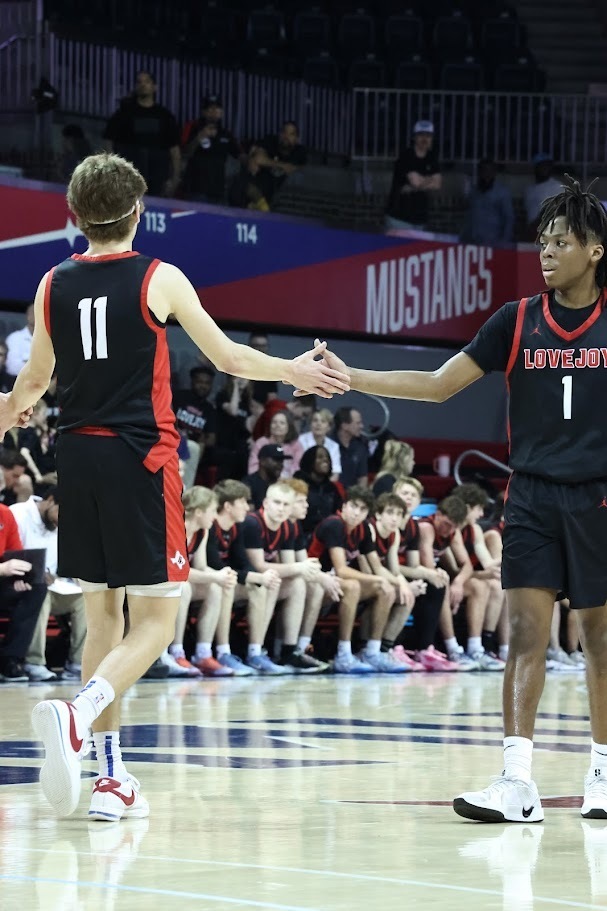 Two basketball players in uniform, one wearing the number 11 and the other 1, shake hands on a court. Behind them, spectators watch from the stands.