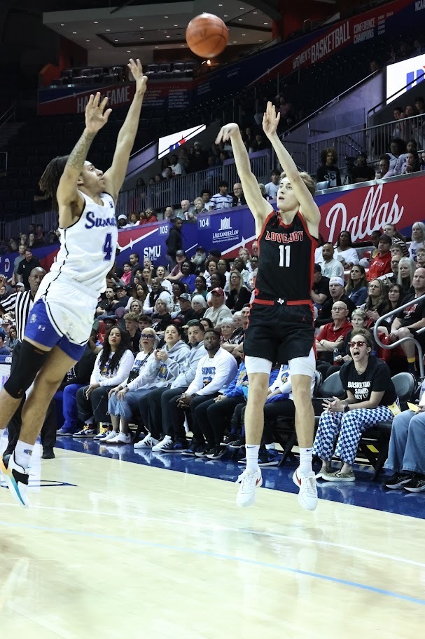 Two basketball players leap in the air; one is reaching for the ball, and the other blocks the shot. Spectators watch from the stands.