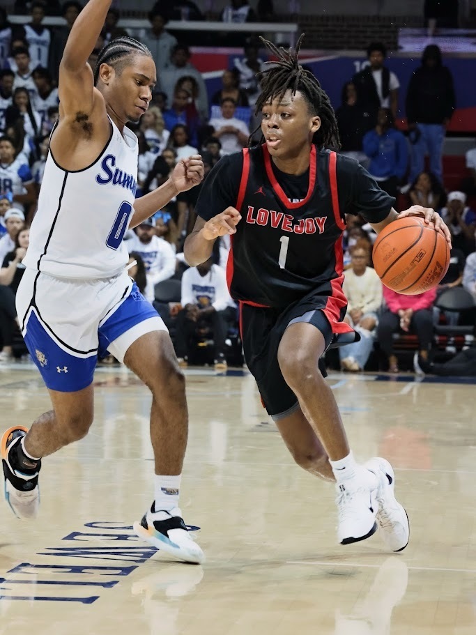 Two basketball players on a court. One wears a white jersey with blue shorts, the other a black jersey with red accents. The player in black dribbles the ball. Audience in the background.