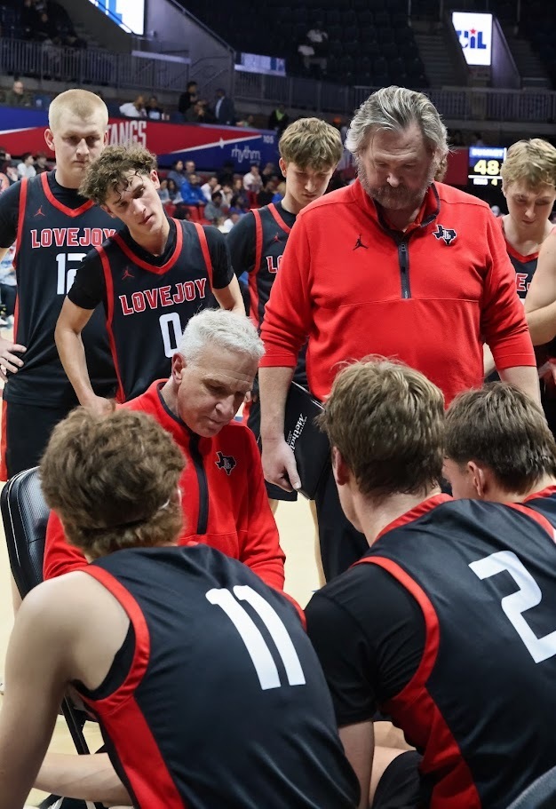 A basketball coach in a red shirt talks to a team of players wearing black and red jerseys.