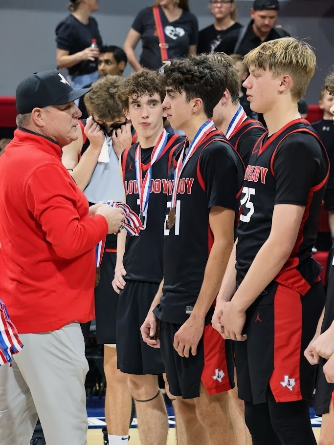 A coach in red hands out medals to four players wearing black jerseys and shorts on a basketball court.