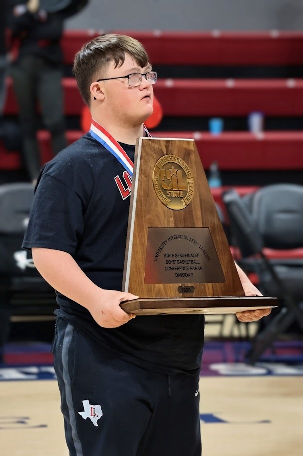 A young man wearing glasses and a medal stands on a basketball court, holding a wooden trophy with a golden emblem.