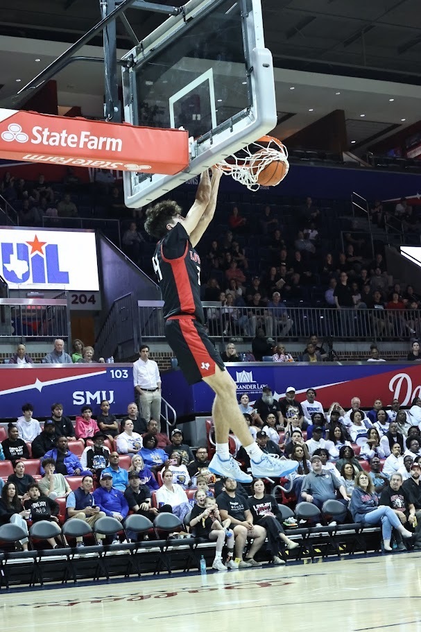 A basketball player dunks a ball. The player wears a red and black jersey. The audience sits in rows of chairs behind him.