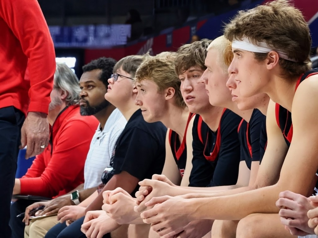 Group of athletes in sports uniforms sit on a bench, watching a game. A man in a red shirt stands nearby.