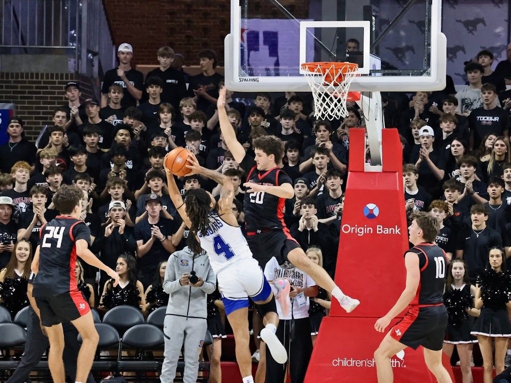 Basketball players compete on a court, with one leaping for a basket. A crowd watches from stands.