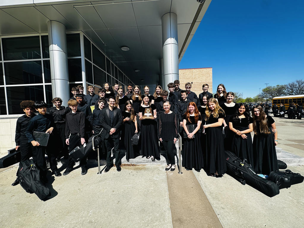 Group of individuals in black attire, likely a choir, posing with instruments and music stands in front of a modern building.
