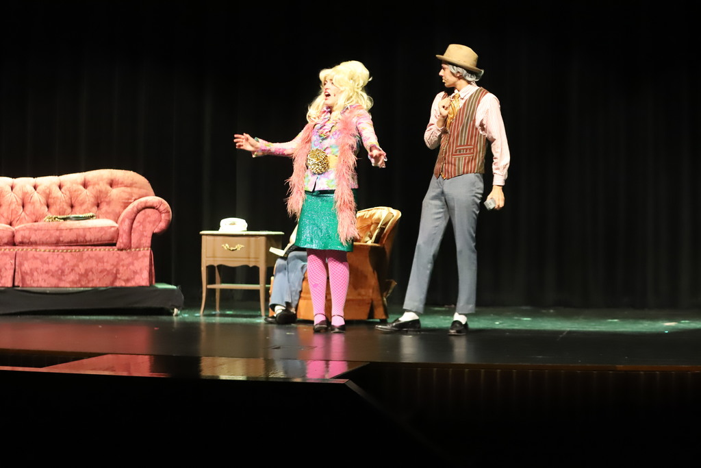 Two individuals on a stage, one in a pink outfit and hat, the other in a white shirt and vest.