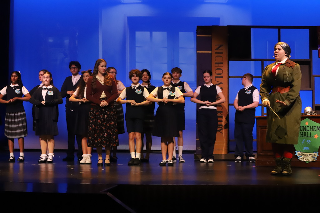 A group of children in uniform stands on stage, with one adult in a green outfit and hat.