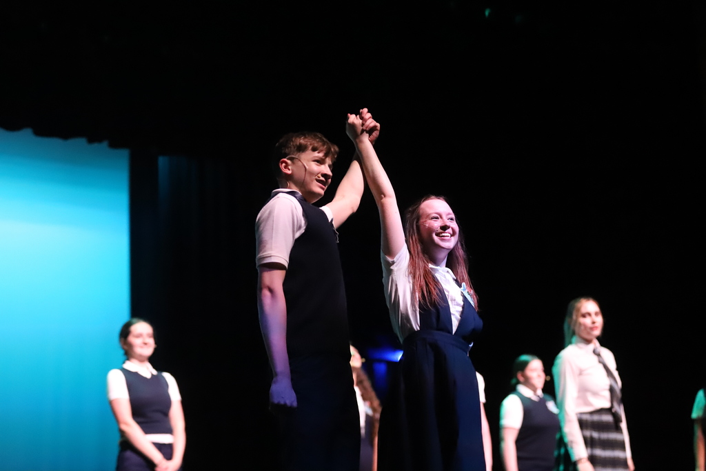 People on stage. Two people hold hands. They smile. Other people in school uniforms behind them.