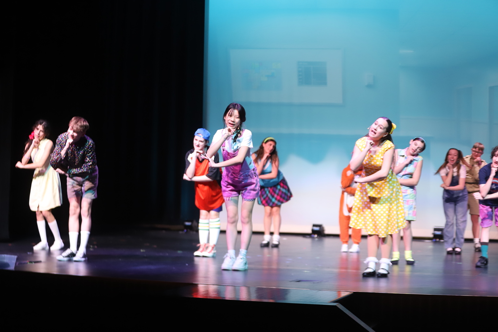 People dressed in various colors perform on stage, with a projector screen in the background.