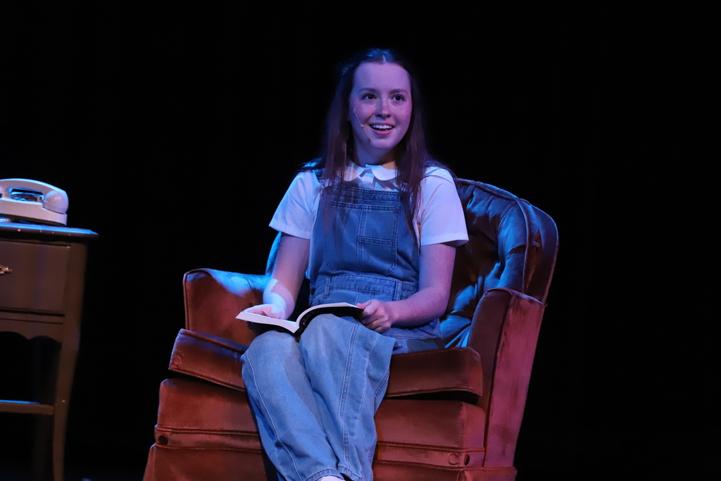 A girl in denim overalls sits in a chair on stage, holding a book and smiling.