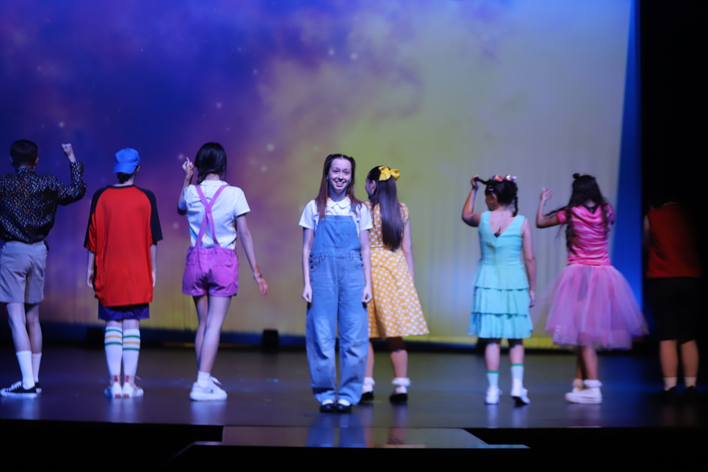 A group of performers on stage, each dressed in different colors, standing in front of a large screen.