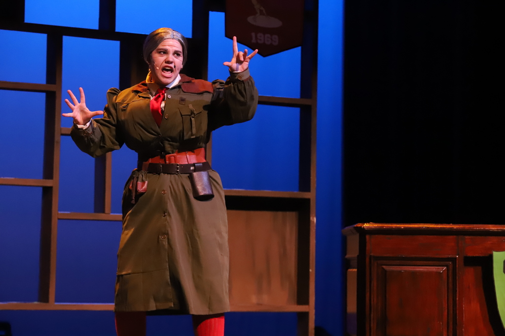 A person in a green outfit on a stage, gesturing with hands while a wooden podium and blue backdrop are behind.
