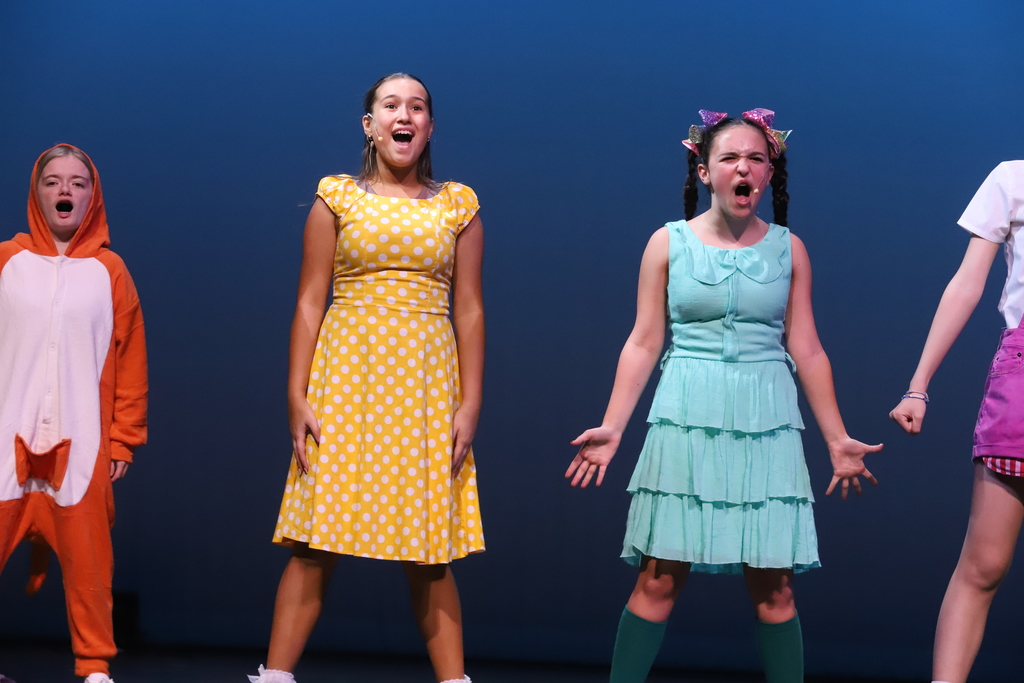 Four performers on stage, with one in a yellow polka-dot dress, another in orange, and two in blue and pink.