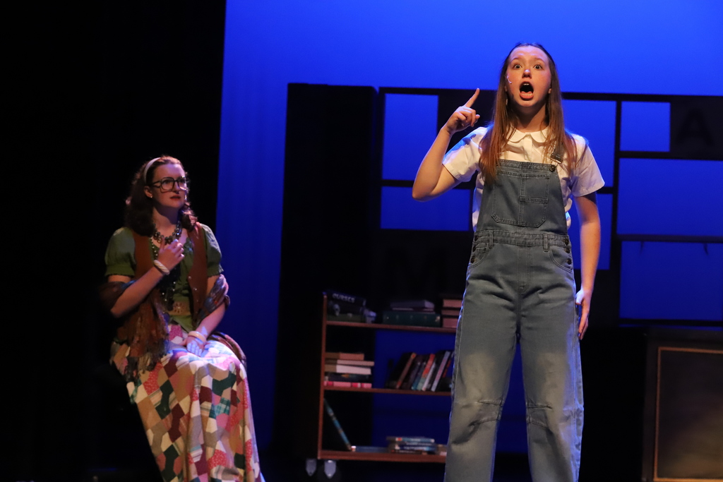 Two women on a stage. One standing with arms raised, another seated. Bookshelf behind them.