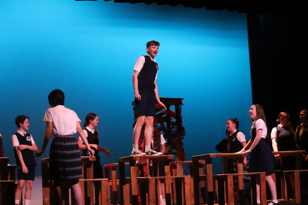 A person stands atop a wooden structure as others perform around them on stage with a blue background.